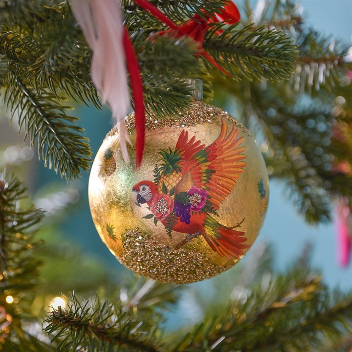 A round gold Christmas ornament decorated with a colourful parrot and glitter, hanging from a green Christmas tree branch with a red ribbon.
