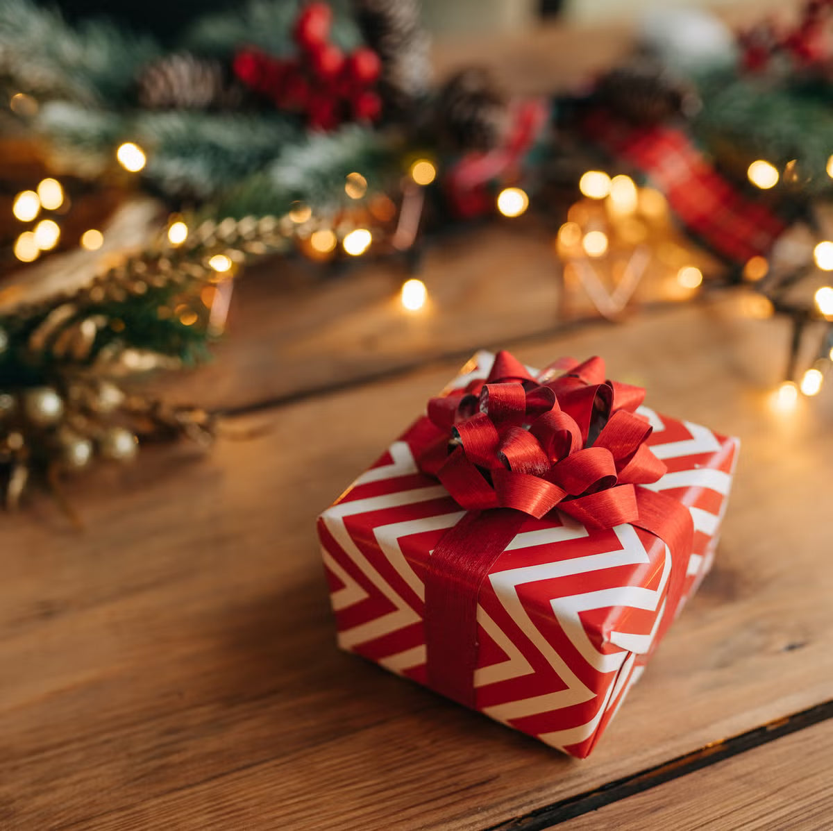 Gift box with red and white pattern and bow on a wooden surface with Christmas decorations in the background.